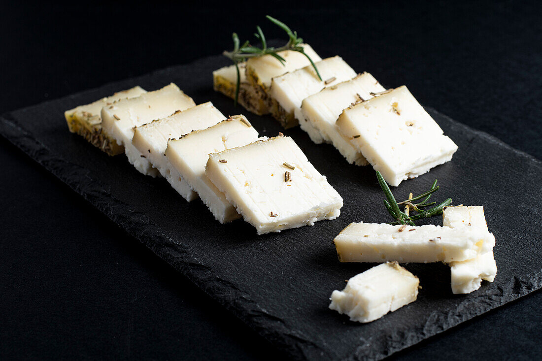 Appetitanregender frischer Schnittkäse auf einem Brett mit Rosmarinzweigen vor schwarzem Hintergrund