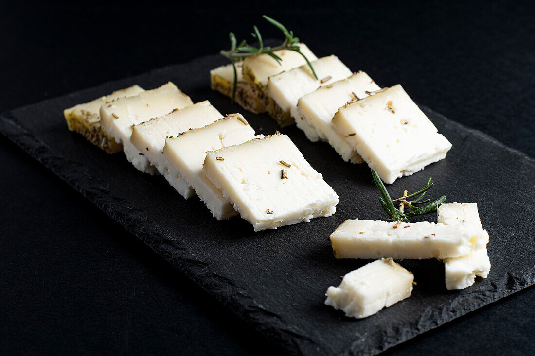 Appetizing fresh sliced cheese placed on board with rosemary sprigs against black background