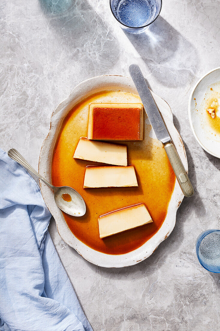 Top view of tasty homemade flan Spanish dessert topped with caramel sauce served on big plate with spoon and knife placed on marble table