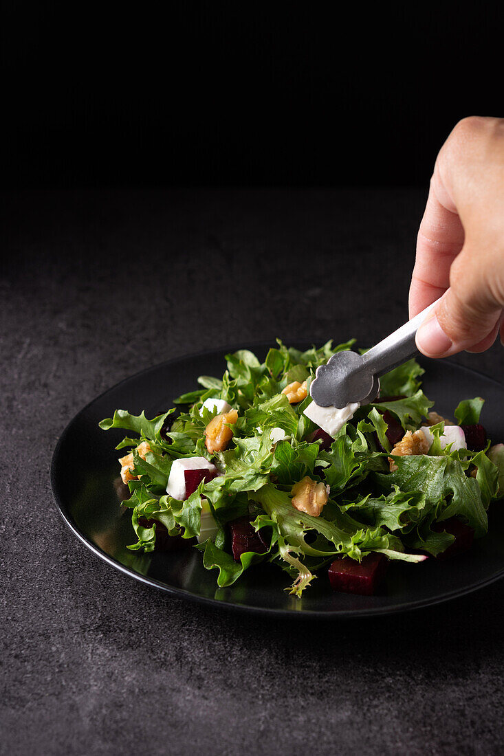 Crop anonymous cook with kitchen tongs serving appetizing beetroot salad with Tofu cheese and lettuce leaves on plate on dark table in studio