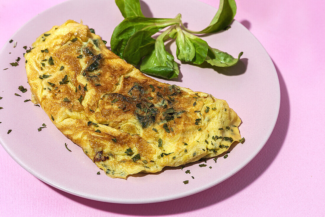 Tasty omelette on plate against fresh parsley sprigs with garlic cloves on pink background