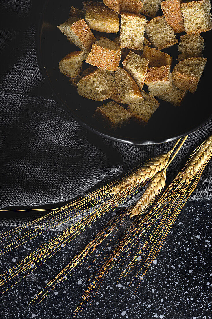 Knusprige Brotstücke in einer Schüssel neben Weizenspitzen auf schwarzem Stoff im Zimmer