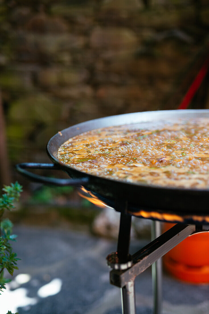 Große Eisenpfanne mit dampfenden Zutaten auf dem Feuer bei der Zubereitung einer traditionellen spanischen Paella im Garten