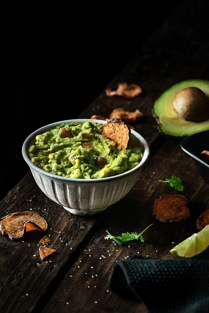 Top view of bowl of fresh guacamole placed near avocado and chips on weathered lumber tabletop
