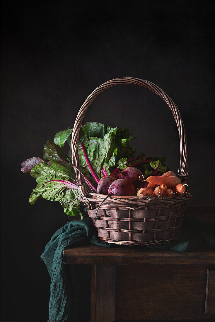 Stilleben eines Korbes mit Gemüse auf einem Holztisch vor schwarzem Hintergrund