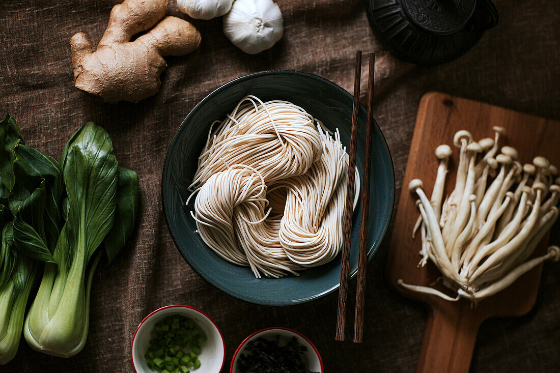 Draufsicht auf verschiedene Zutaten für eine leckere Ramen-Zubereitung auf einem braunen Tischtuch in der Küche