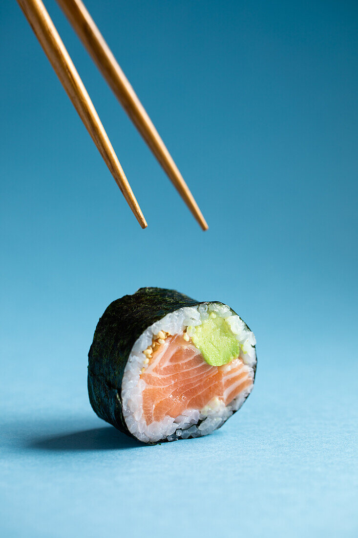 Tasty Japanese Futomaki Norwegian sushi roll with fresh salmon and avocado placed against blue background in studio with wooden chopsticks