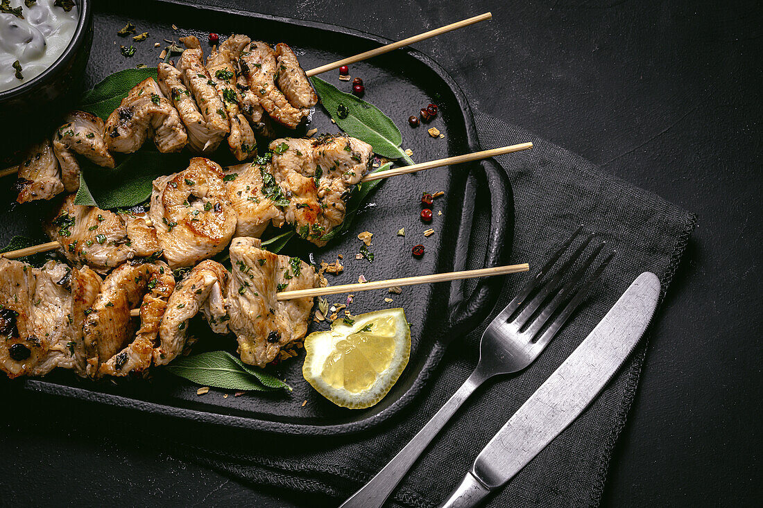 From above of appetizing freshly cooked meat on skewers served on tray on black table with bowl of sauce