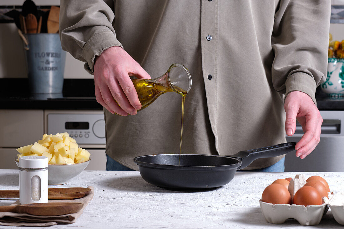 Anonyme Person gießt Olivenöl in eine Bratpfanne, während sie ein hausgemachtes spanisches Omelett mit Eiern und gehackten Kartoffeln auf einem Tisch in einer Küche zu Hause zubereitet