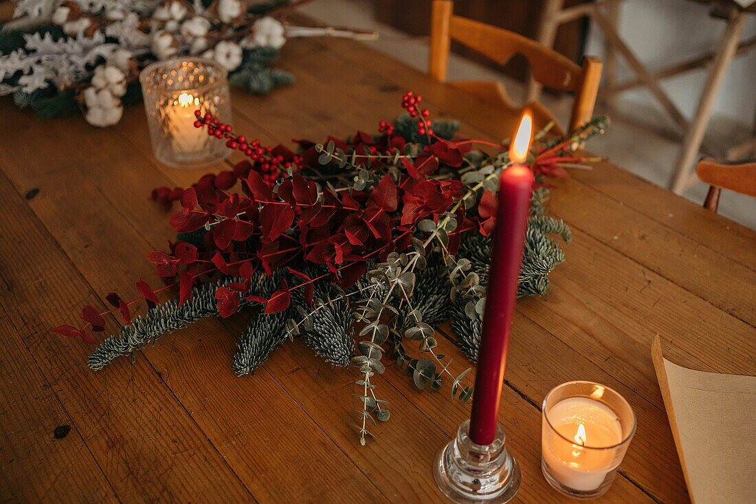 From above of festive Christmas bouquet with branches of cotton, fir and twigs of eucalyptus and bright red branches with berries placed on wooden table with candles in room