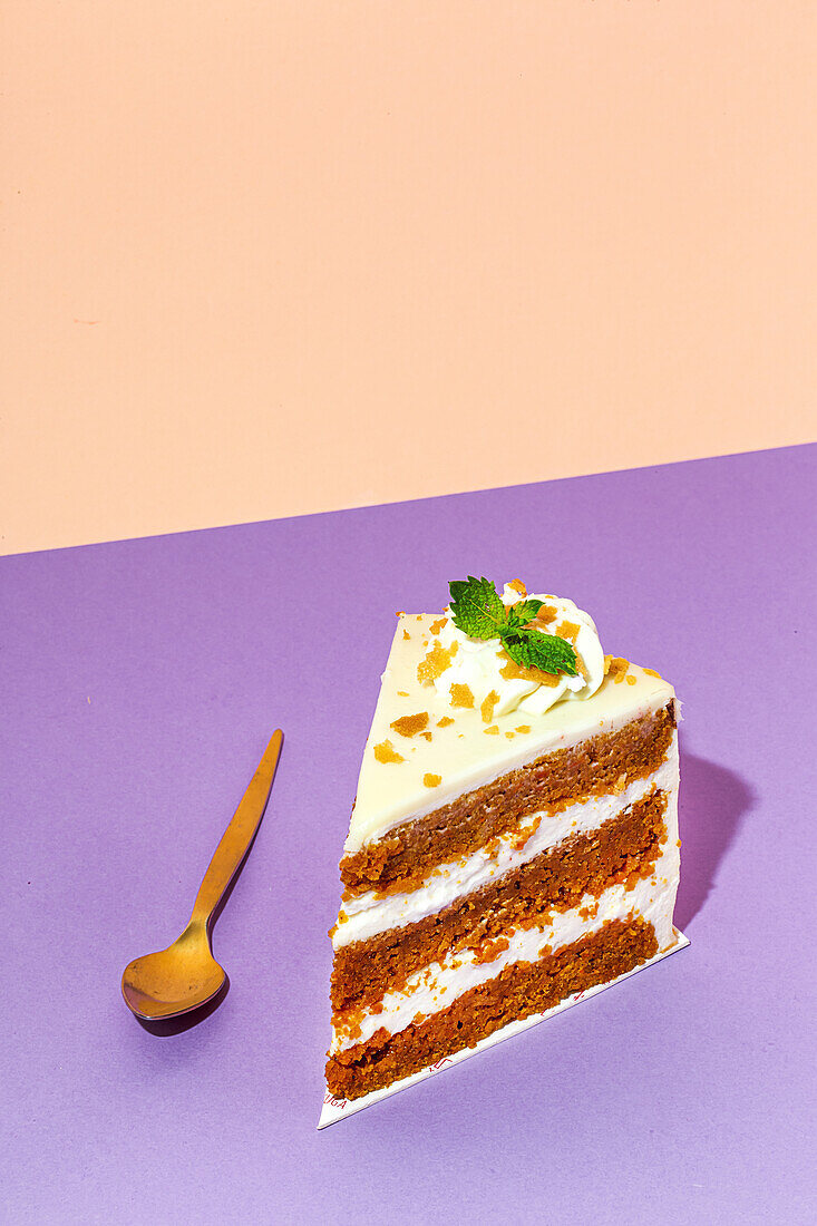 Slice of tasty sweet carrot sponge cake with cream decorated with mint leaf served on plate with spoon on table on colorful background