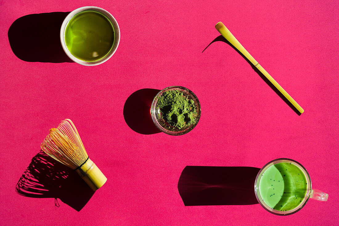 Blick von oben auf einen hellen Hintergrund mit einem Glas grünem Tee und getrocknetem Matcha mit Bambus-Chasen und Chashaku auf einer rosa Fläche