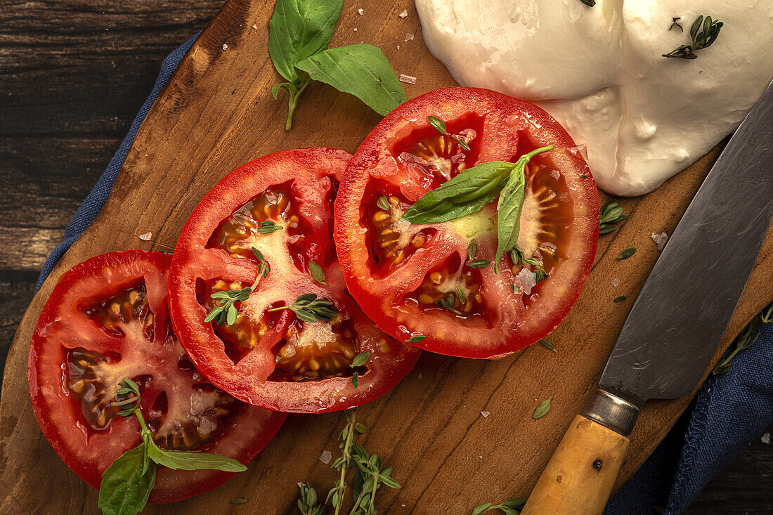 Draufsicht auf köstlichen Mozzarella und Scheiben frischer Tomaten mit grünen Basilikumblättern und Thymian, serviert auf einem hölzernen Schneidebrett mit Messer auf dem Tisch