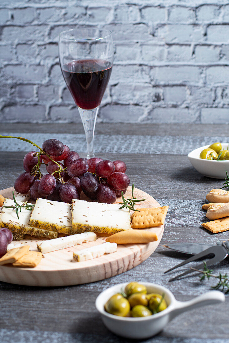 Glass of red wine placed near assorted cheese and grape with crackers and olives decorated with rosemary on table