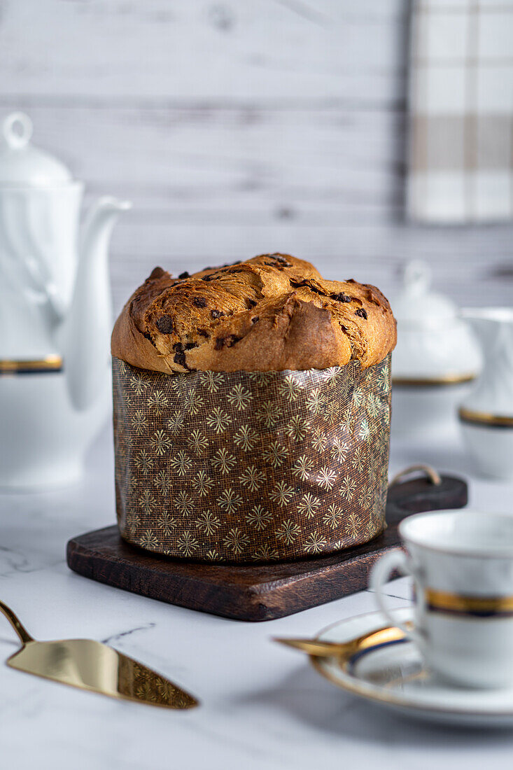 Rustikaler traditioneller Weihnachts-Panettone-Kuchen mit Rosinen auf Holzbrett zwischen luxuriösem Frühstücksgeschirr