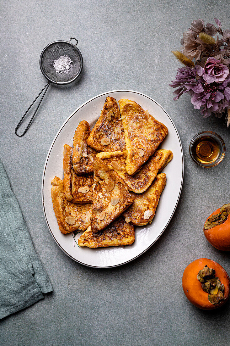 Top view of white plate with delicious French toasts placed near jar with honey , fresh persimmons and sieve with icing sugar â€‹on gray table