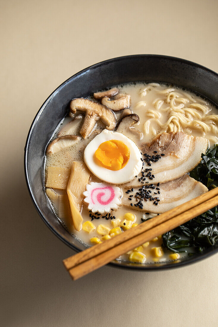 Appetitanregende japanische Ramen mit gekochtem Ei und Pilzen, serviert in einer Schüssel mit Holzstäbchen vor beigem Hintergrund