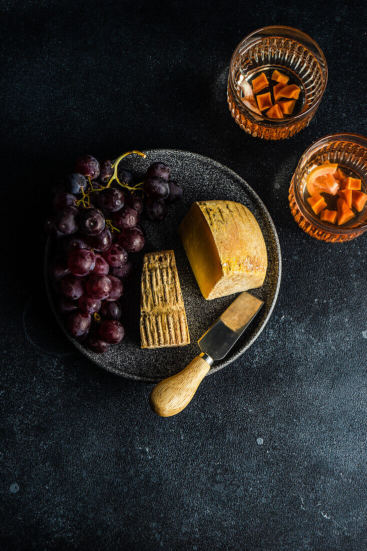 Schwarzer Steinteller mit Käse, rohen leckeren Trauben und Weingläsern auf dunklem Betontisch