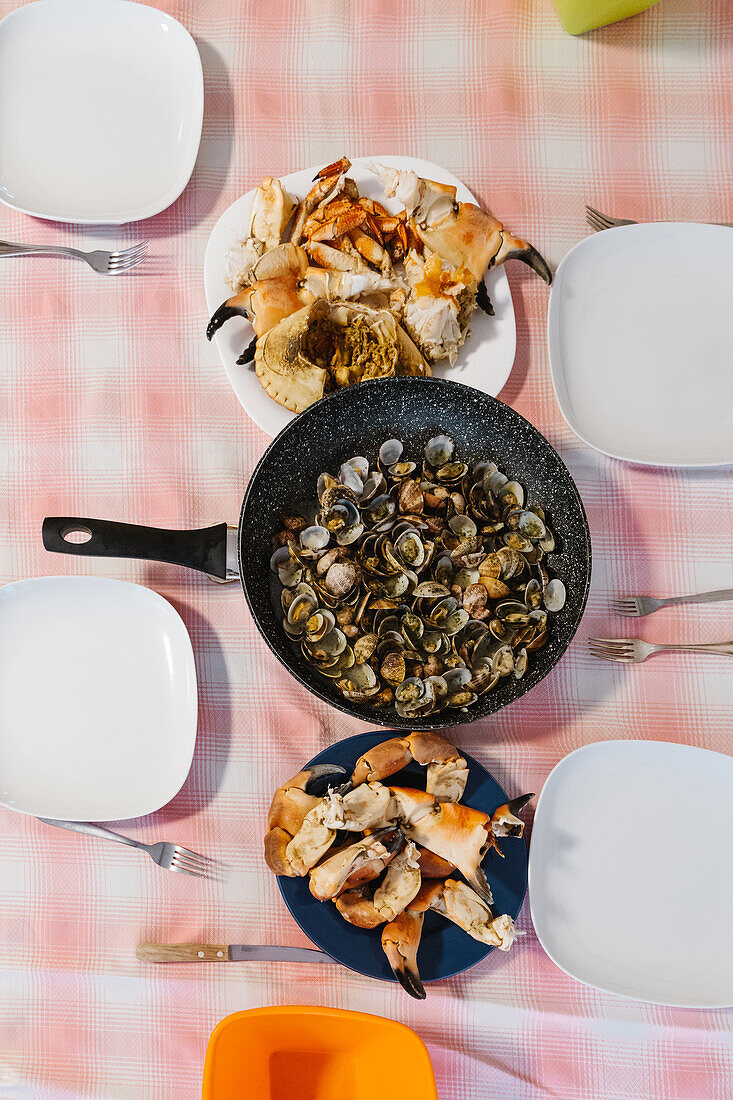 Blick von oben auf eine Bratpfanne mit gebratenen Muscheln, die auf einem Tisch mit Krabbenscheren und weißen Tellern in einer hellen Küche serviert werden