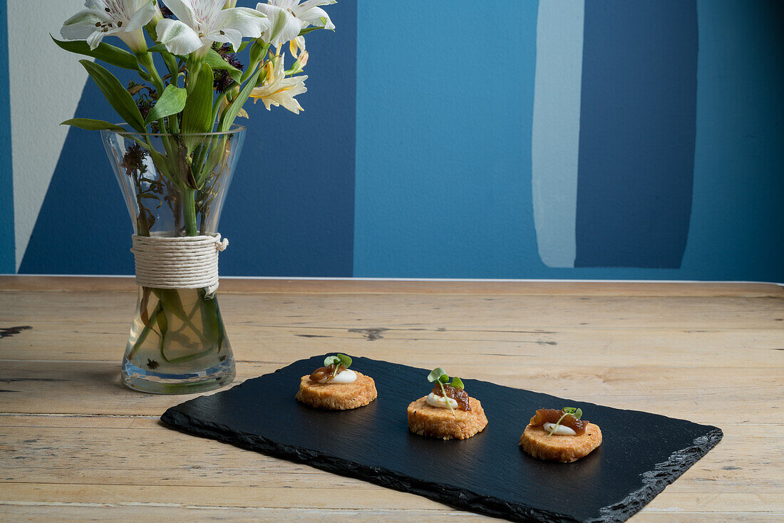 Einzelnes Stück köstlicher gekochter Vorspeisenhappen, serviert auf schwarzem Brett in der Nähe blühender weißer Lilien an blauer Wand