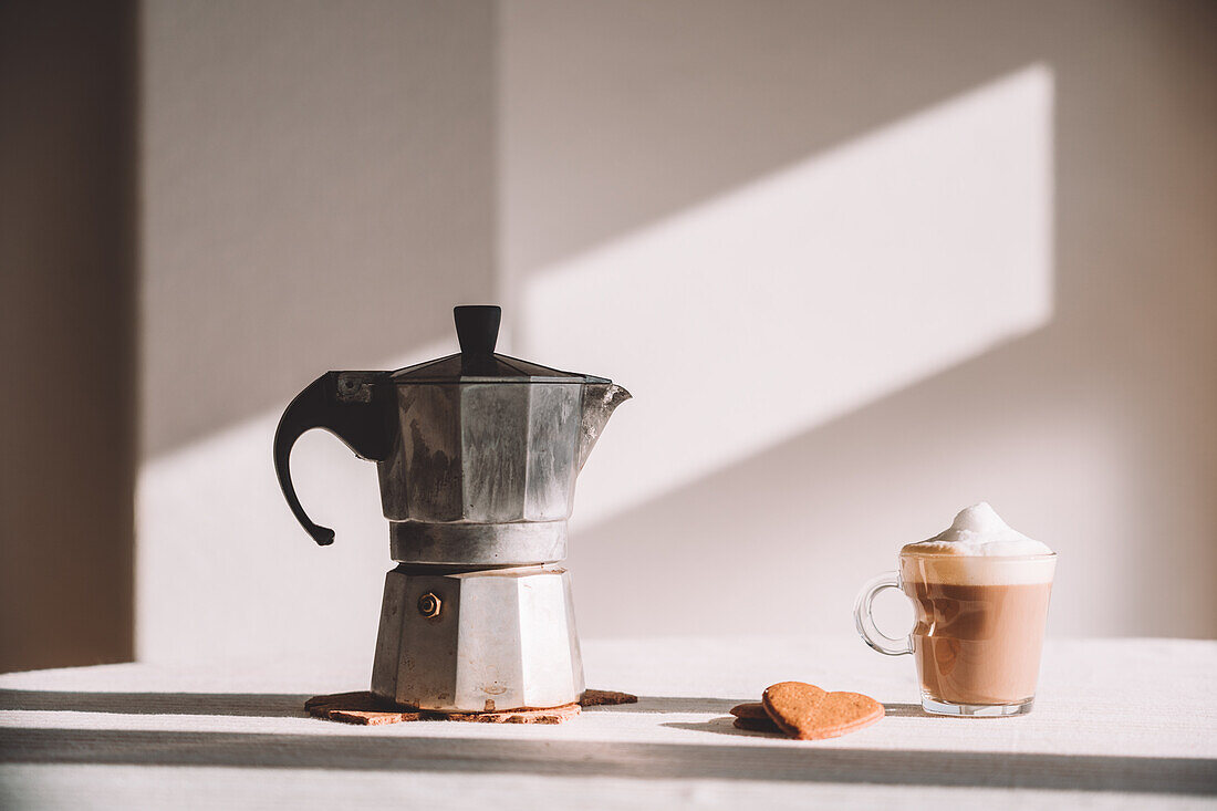 Geysir-Kaffeemaschine aus Metall auf dem Tisch mit aromatischem, schaumigem Latte Macchiato im irischen Glas mit herzförmigen Keksen in der Küche an einer weißen Wand an einem sonnigen Tag