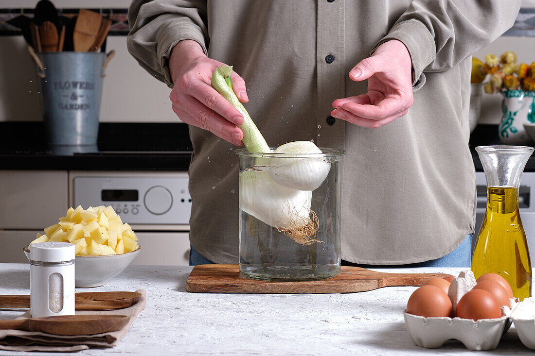 Anonyme Person gießt frische, geschälte Zwiebeln in einen Glastopf mit Wasser, während sie ein hausgemachtes spanisches Omelett mit Eiern und Kartoffeln zubereitet