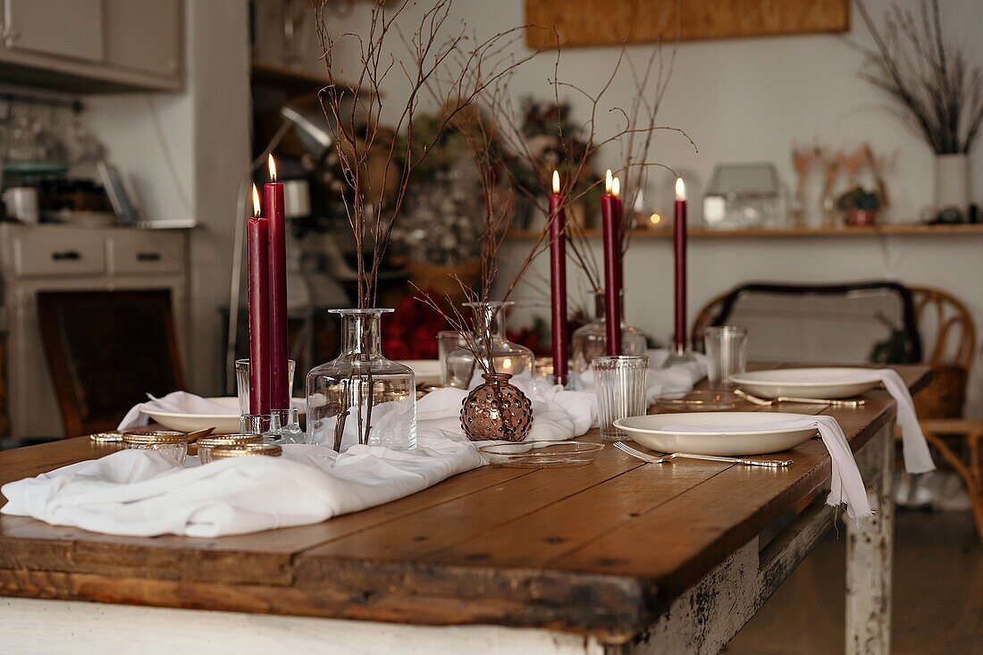 White tablecloth and plates placed on festive table decorated with burning candles and dry branches of tree