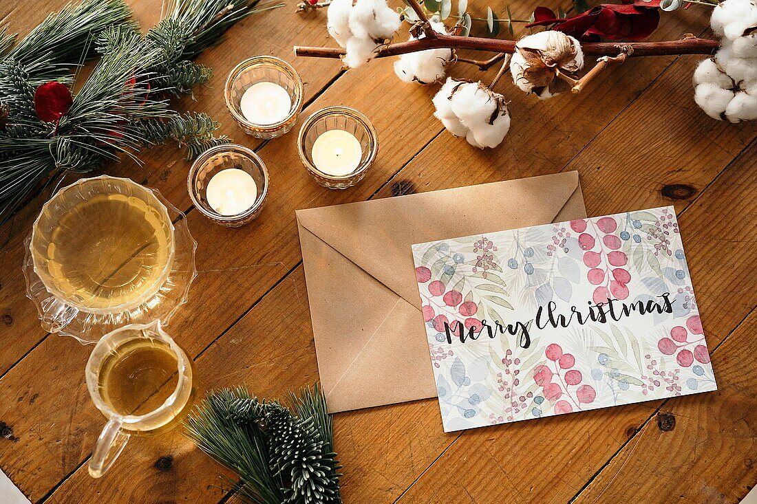 Draufsicht auf eine weihnachtliche Komposition mit einer bunten Postkarte mit der Aufschrift Feliz Navidad in der Nähe von brennenden Kerzen und Teetassen auf einem Holztisch, der mit bunten Pflanzenzweigen dekoriert ist
