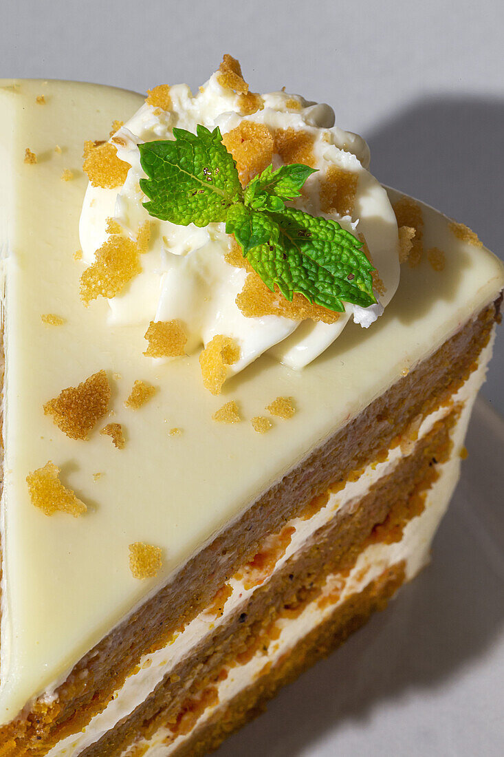 From above close up of slice of tasty sweet carrot sponge cake with cream decorated with mint leaf served on plate with spoon on table on white background