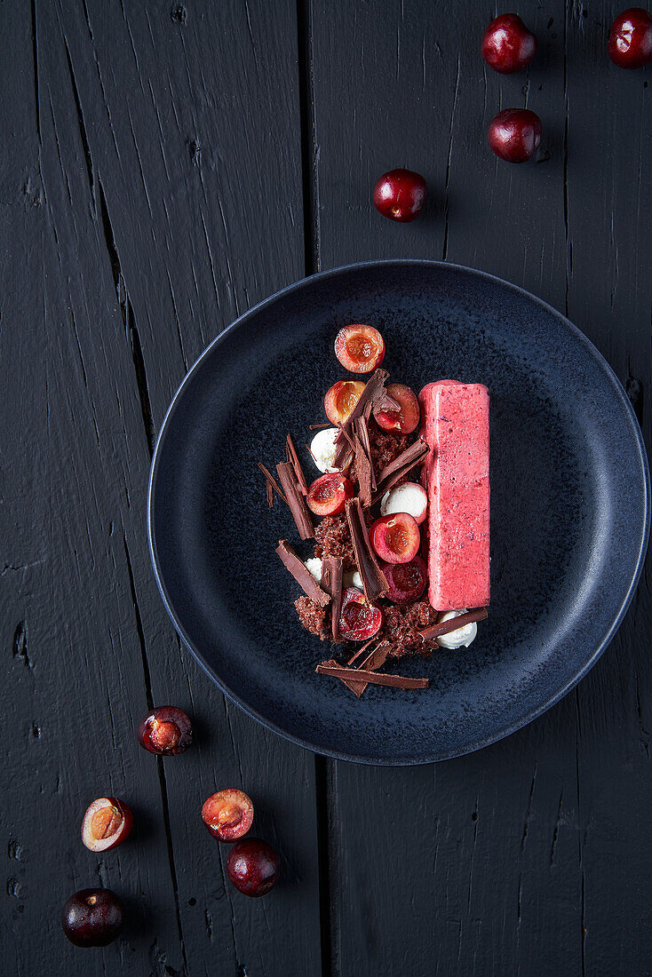 Draufsicht auf eine leckere Beeren-Mousse-Torte, die auf einem schwarzen Teller mit Kirschen und Schokoladenflocken serviert wird und auf einem Holztisch steht