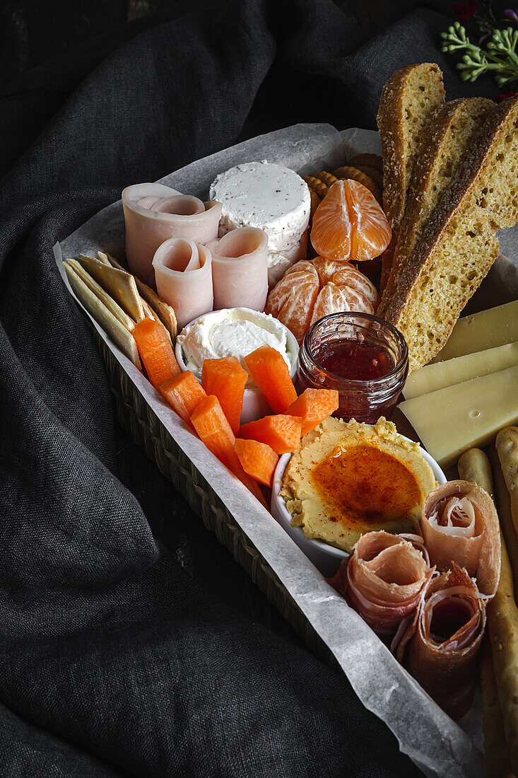 Von oben Brunch-Box mit verschiedenen Wurst- und Käsesorten und Knäckebrot in der Nähe von reifen Kiwis, süßen Erdbeeren und geschälten Mandarinen in der Nähe von Marmelade in einem Glas auf einem Holztisch in der Nähe einer Serviette