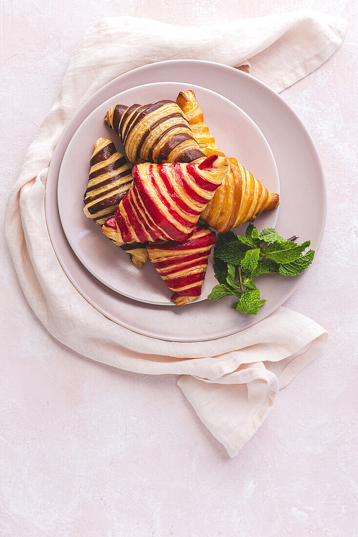 Draufsicht auf leckere Croissants auf einem Teller mit einem Minzzweig auf pastellrosa Hintergrund