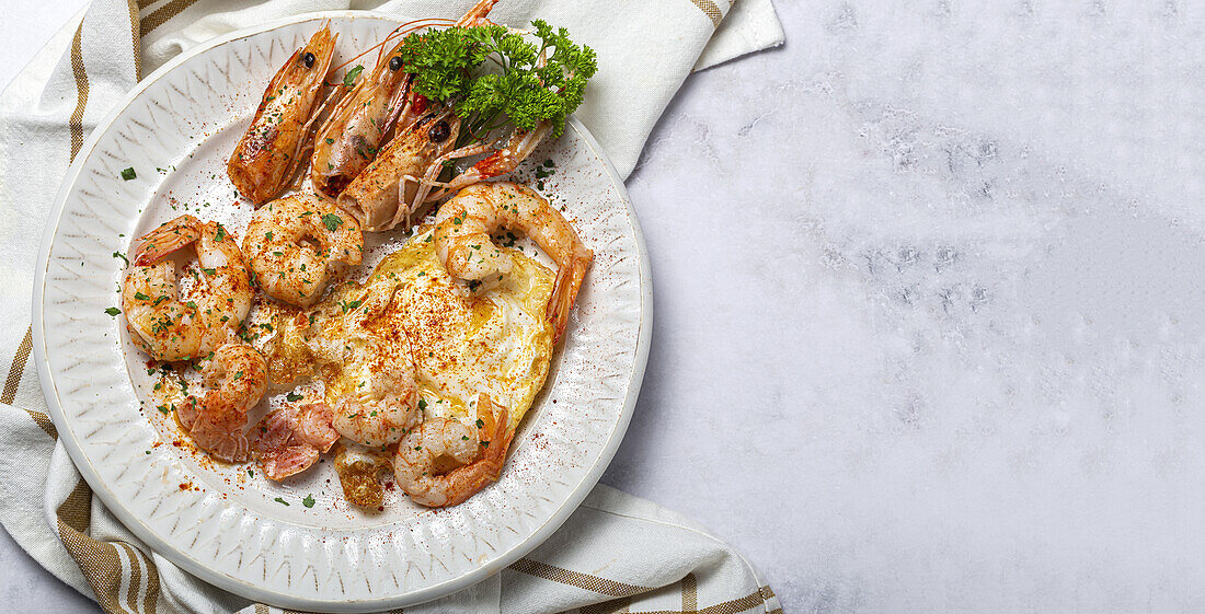 From above appetizing fried eggs with king prawns served on white plate with green herbs and seasoning on towel in light kitchen