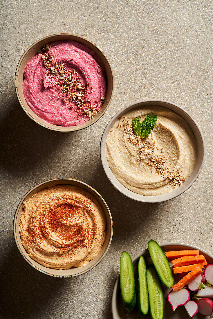 Top view of bowls with assorted hummus served on table with fresh cucumbers and radish