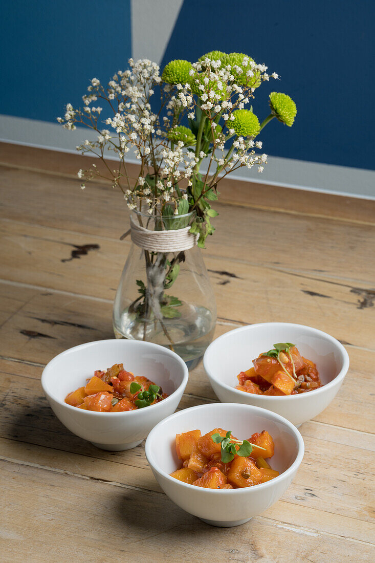 High angle of vegetable stew with pumpkin served in white ceramic bowls placed on wooden floor near blooming gypsophila