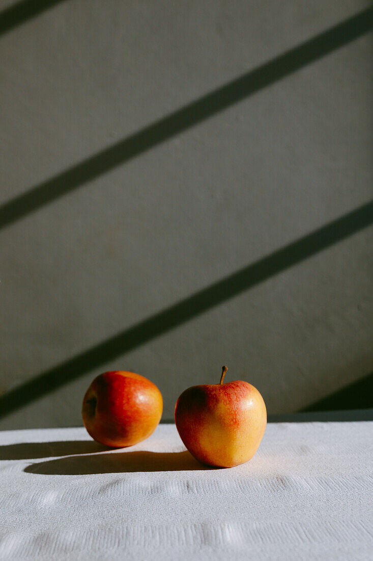 Frische ganze Äpfel auf weißem Tischtuch vor grauer Wand mit Schatten im Tageslicht