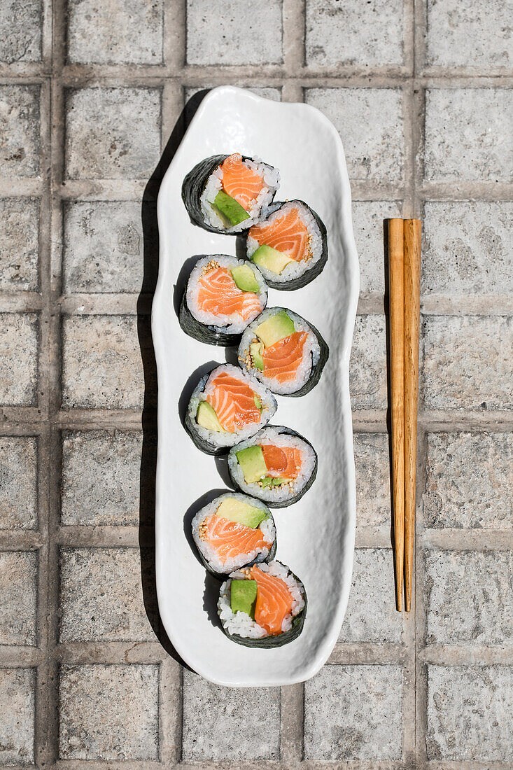 Draufsicht auf leckere japanische Futomaki-Sushi-Rollen aus norwegischem Lachs, die mit Stäbchen auf einem gepflasterten Gehweg in einer sonnigen Straße in der Stadt platziert werden