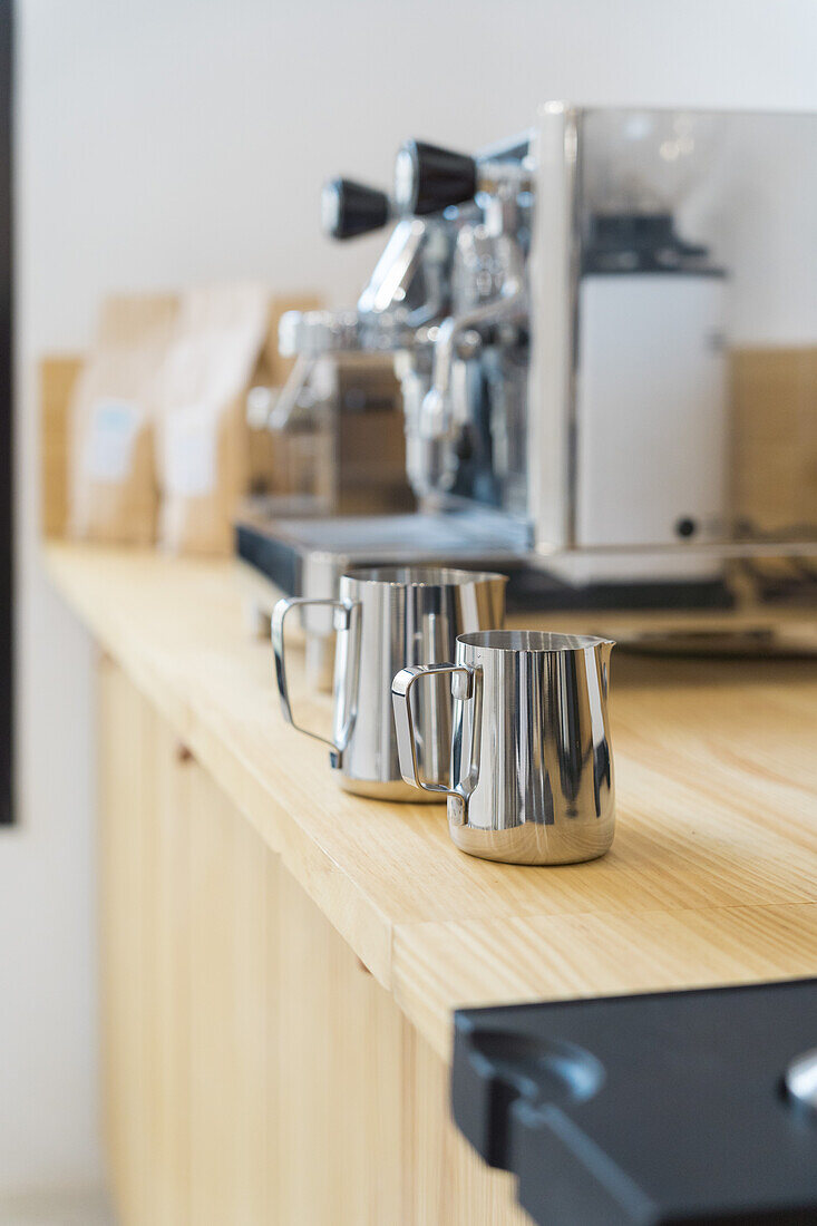 Soft focus of stainless professional pitchers for pouring milk placed on wooden counter in modern coffee house with coffeemaker machines