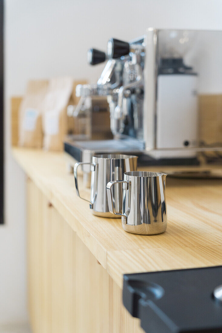 Weichzeichner von rostfreien professionellen Krügen zum Ausgießen von Milch auf einer Holztheke in einem modernen Kaffeehaus mit Kaffeemaschinen