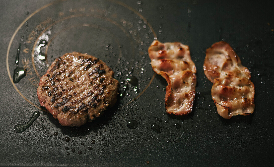 From above grilled patty for burger and bacon slices on black stove