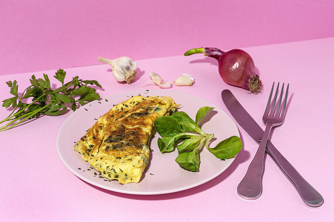 Tasty omelette on plate against fresh parsley sprigs and red onion with garlic cloves on pink background