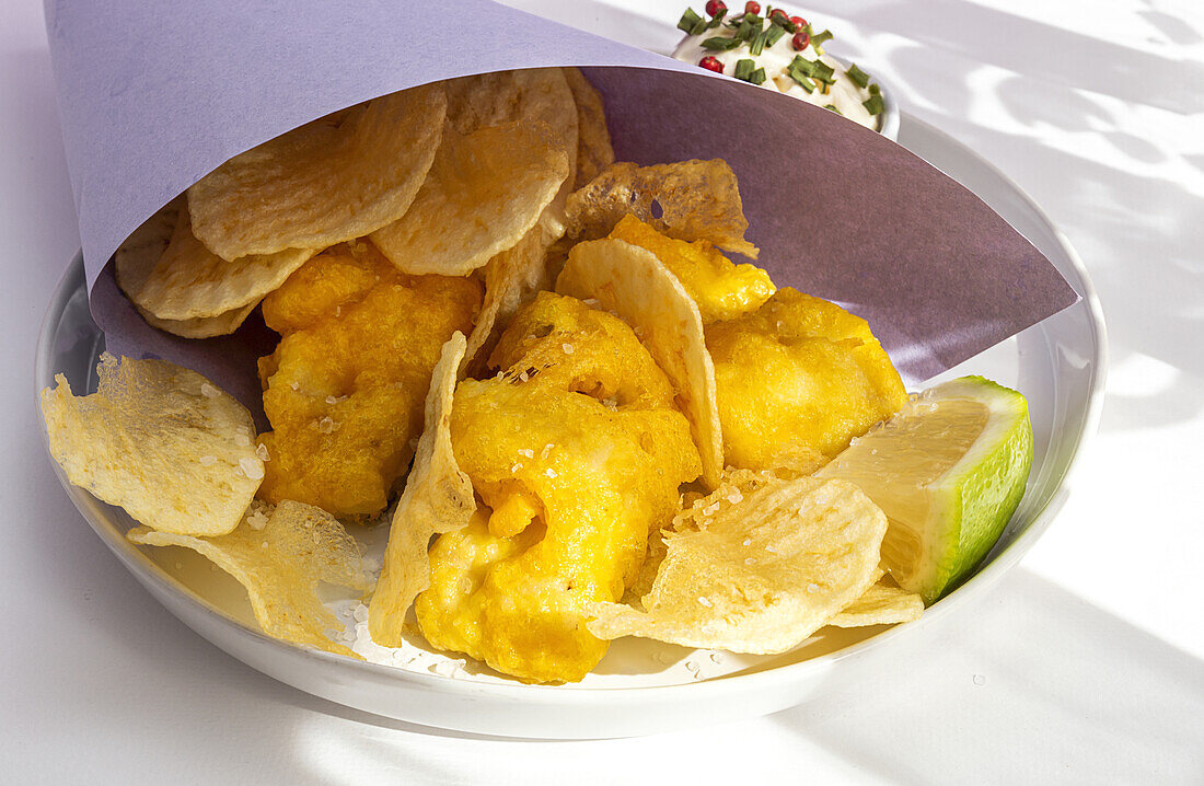 Appetizing fried chips and fish in paper wrapping served on plate with sauce on table with bright sunlight in cafe