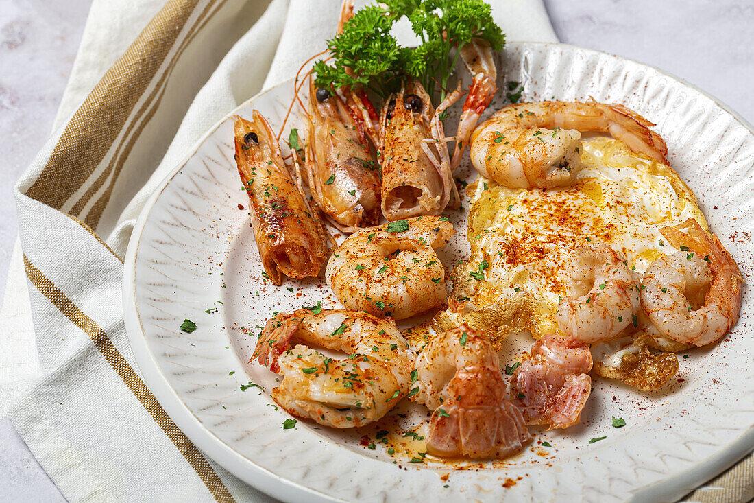 From above appetizing fried eggs with king prawns served on white plate with green herbs and seasoning on towel in light kitchen