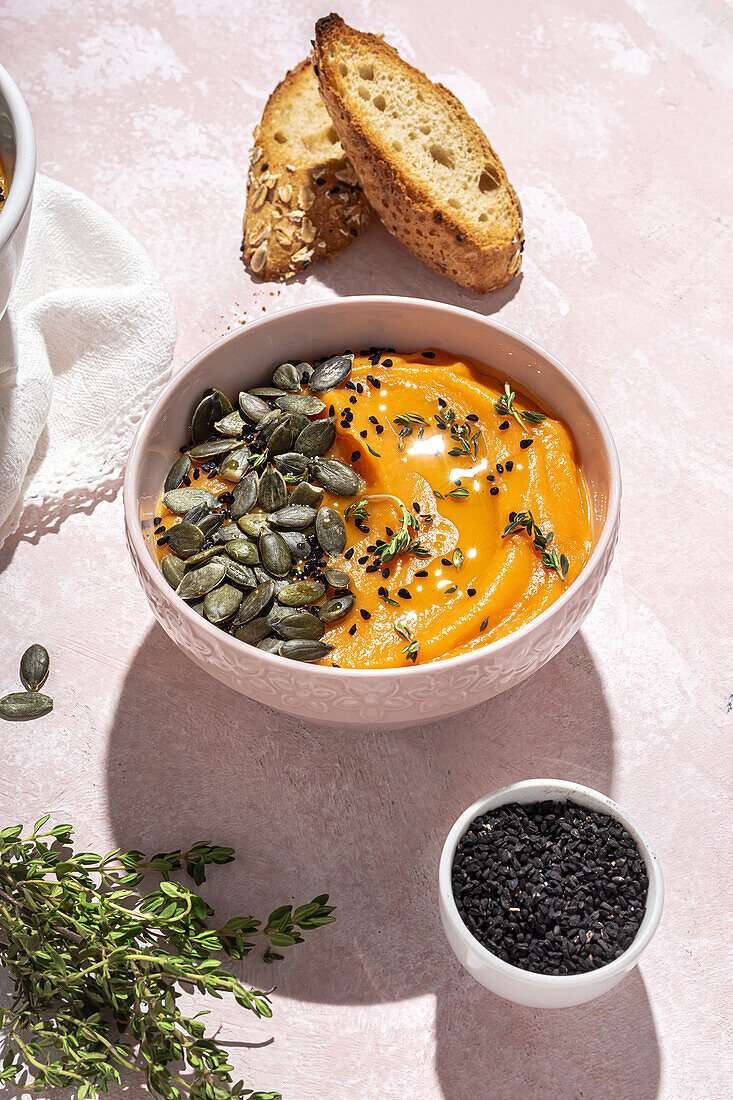 Appetitliches hausgemachtes Kürbispüree mit Kernen und Kräutern in einer Schüssel auf einem Tisch neben knusprigen Brotscheiben in der Küche von oben