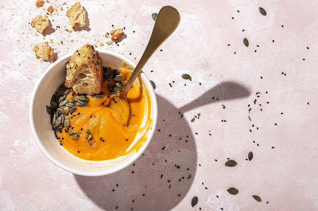 From above of spoon in white ceramic bowl with eaten pumpkin cream soup placed on table near scattered seeds and crispy bread pieces