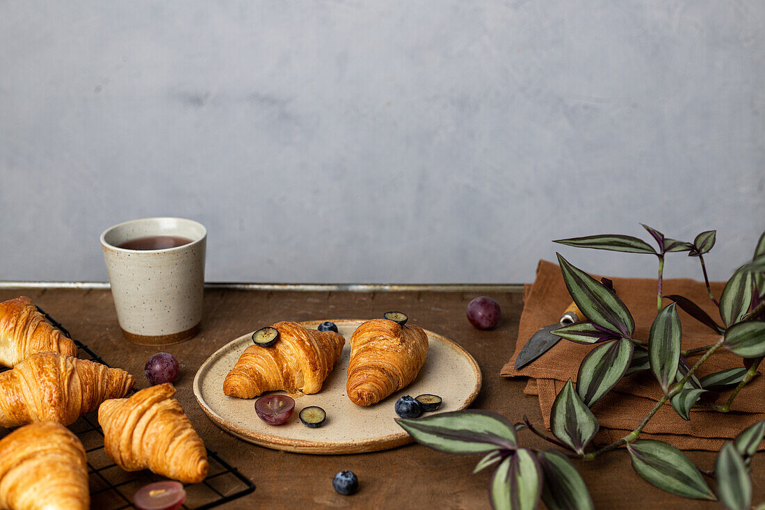Leckere frisch gebackene Croissants auf einem Teller mit Früchten neben einer Tasse Tee auf einem Holztisch am Morgen in einem hellen Raum