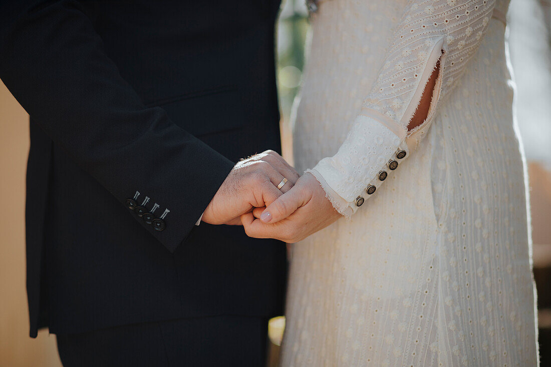 Seitenansicht eines anonymen Bräutigams, der die Hände einer eleganten Braut im weißen Hochzeitskleid hält