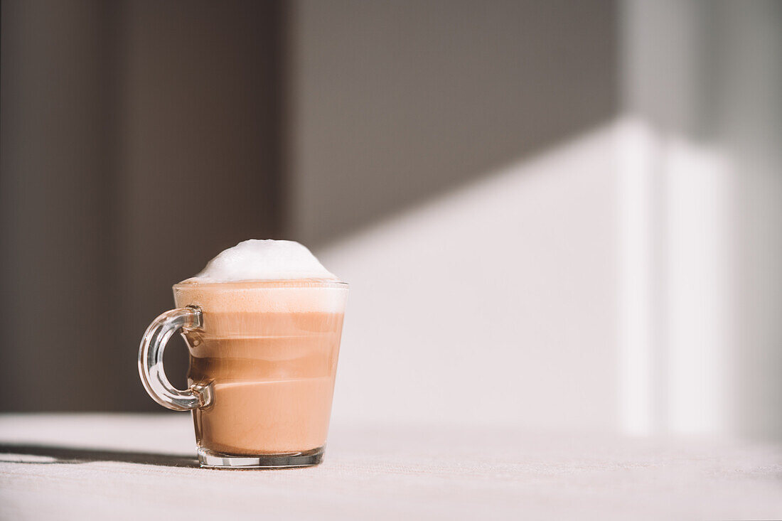 Glass of foamy latte macchiato served on table on sunny day in kitchen