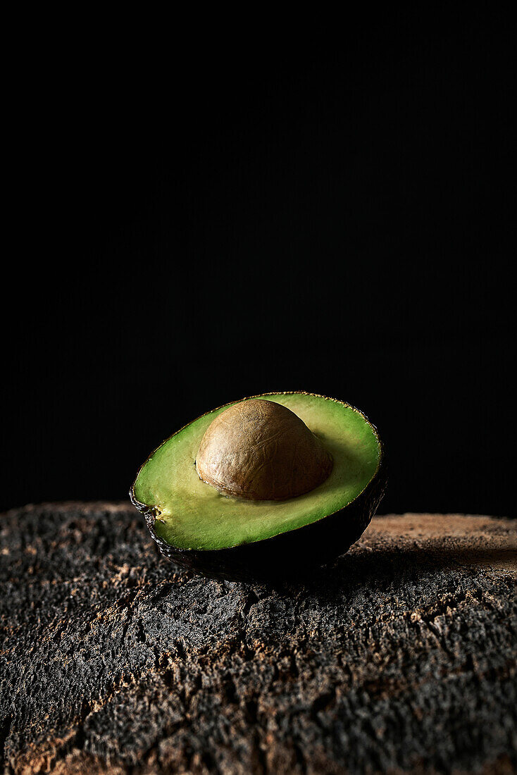 Half of ripe avocado with seed placed on rough surface against black background