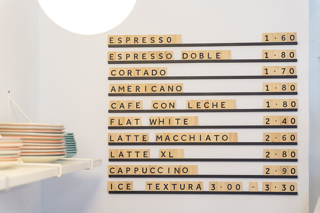 Menütafel mit verschiedenen Kaffeegetränken und Preisen an einer weißen Wand neben einem Tresen mit Geschirr in einem modernen hellen Kaffeehaus mit strahlendem Licht